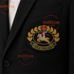 Op de afbeelding: Een zwarte blazer met een wit overhemd eronder en een opvallende geborduurde patch op de borst. De patch toont een ridder te paard, een schild met de letter B en de tekst BURBERRYS omringd door gouden korenaren op een zwarte achtergrond.