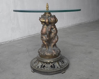 May include: A bronze-toned side table featuring a round glass top. The base is adorned with sculpted cherub figures and rests on a decorative, circular base. The table's design is ornate and vintage.