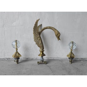 Puede incluir: Un grifo de cisne de latón vintage con un pomo de bola de cristal a cada lado. El grifo de cisne tiene alas detalladas y un cuello curvado. Los pomos de bola de cristal están montados sobre bases de latón. El grifo y los pomos están sobre un fondo neutro.