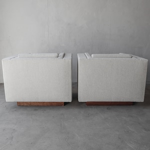 May include: Two white upholstered chairs with wooden bases. The chairs are facing away from the camera and are positioned side by side.