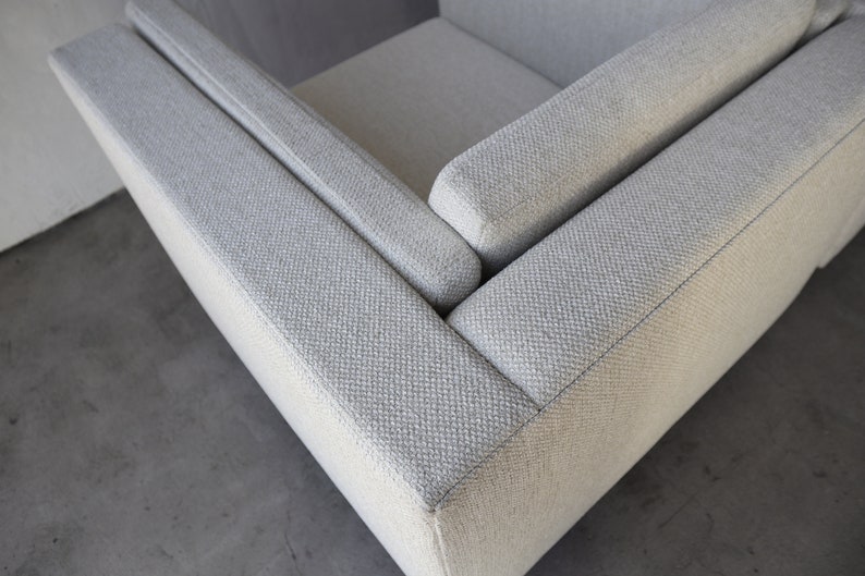 May include: Close-up of the corner of a light gray fabric sofa with a textured weave. The sofa has a square armrest and a thick, padded cushion.