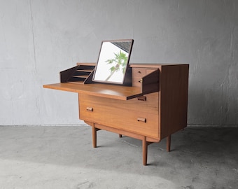 Könnte beinhalten: Ein Mid-Century Modern Schreibtisch aus Holz mit eingebautem Spiegel. Der Schreibtisch hat eine rechteckige Form mit Schubladen und einer herunterklappbaren Schreibfläche. Der Spiegel ist rechteckig und angewinkelt. Der Schreibtisch ist aus hellbraunem Holz.