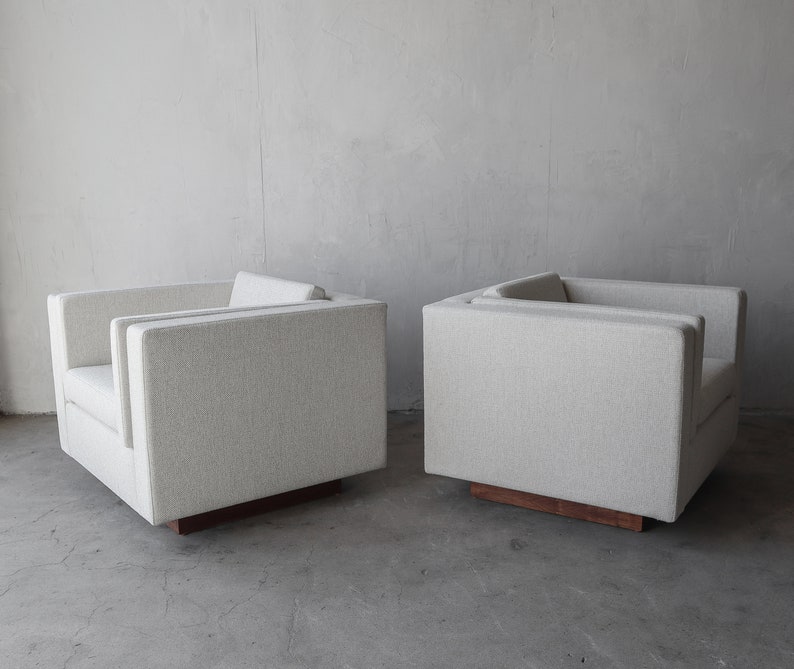 May include: Two white upholstered armchairs with wooden bases. The chairs are positioned side by side on a gray concrete floor.