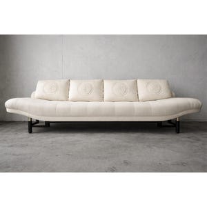 May include: Cream-colored sofa with four decorative pillows. The sofa has a curved design with a tufted seat and back. The pillows feature a circular embossed pattern. The sofa rests on black legs with gold accents, offering a modern aesthetic.