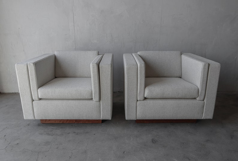 May include: Two light gray upholstered armchairs with a wood base. The chairs have a simple, modern design.