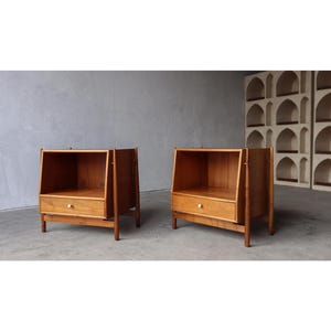 May include: Two wooden nightstands with a mid-century modern design. Each nightstand has a drawer with a white knob and an open shelf. The wood has a warm, natural finish. The nightstands are set against a gray wall.