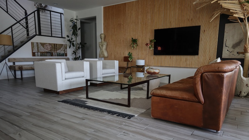 May include: A living room with a brown leather sofa, two white armchairs, a glass coffee table, and a wooden wall with a flat screen TV.