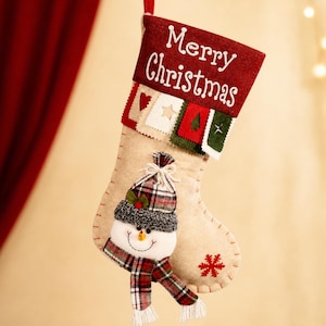 May include: A beige felt Christmas stocking featuring a red cuff with the white text Merry Christmas. Below the cuff are small fabric squares with heart, star, and tree motifs. A plush snowman with a plaid hat and scarf is attached to the front.