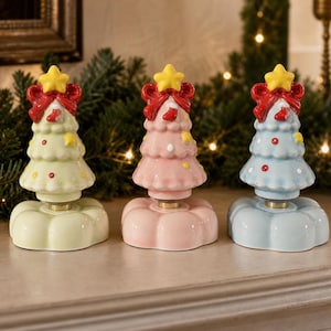 Op de afbeelding: Drie keramische kerstboombeeldjes in pastelgroen, roze en blauw. Elke boom heeft een gele ster als piek, een rode strik, een klein sokje en kleurrijke stippen als versiering, geplaatst op een geschulpte, ronde voet van keramiek.