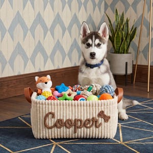 Op de afbeelding: Een beige corduroy hondenmandenmand met bruine leren handvatten en de naam "Cooper" in bruin schrift. De mand is gevuld met kleurrijk hondenspeelgoed en een jonge husky met een blauwe halsband zit erachter.