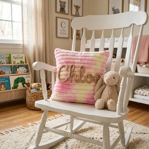 Op de afbeelding: Een witte houten schommelstoel met een vierkant kussen met tie-dye patroon in roze en gele tinten. Het kussen heeft de naam Chloe in reliëf geschreven met beige touw in het midden. Een beige pluche konijn zit op de zitting van de stoel.