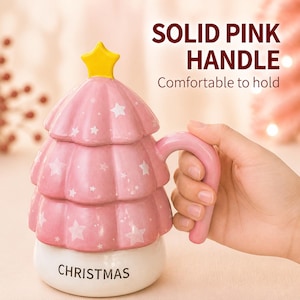 May include: A pink ceramic mug shaped like a Christmas tree with a yellow star topper and a lid. The mug features white star patterns and the word CHRISTMAS printed on the base. It includes a solid pink handle and text reading SOLID PINK HANDLE.