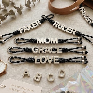 Könnte beinhalten: Anpassbare Namens-Schlüsselanhänger mit silbernen Metallbuchstaben, die "FAKER", "RULER", "MOM", "GRACE" und "LOVE" buchstabieren. Die Schlüsselanhänger haben schwarze und silberne Kordeln. Zusätzliche silberne Buchstaben und ein Etikett sind ebenfalls sichtbar.