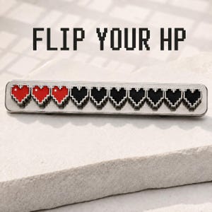 Puede incluir: Un broche rectangular de metal con el texto FLIP YOUR HP en una fuente pixelada. El broche muestra una fila de diez corazones de arte de píxeles, con los tres primeros corazones de color rojo y los siete restantes de color negro, bordeados en plata.