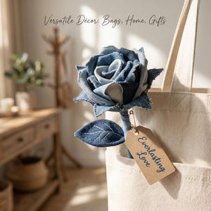 May include: A denim rose flower with frayed edges and a single dark blue leaf on a stem, attached to a beige canvas tote bag with a safety pin. A brown paper tag hangs from the pin with the text Everlasting Love. Text above reads Versatile Décor: Bags, Home, Gifts.