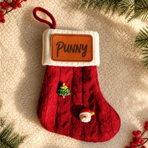 May include: A red cable-knit Christmas stocking with a white ribbed cuff featuring a brown leather patch embossed with the text PUNNY. The stocking is decorated with a small green Christmas tree charm and a miniature Santa Claus face charm.
