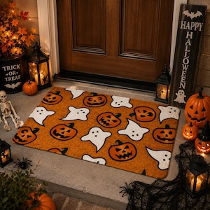 Op de afbeelding: Een rechthoekige deurmat met een oranje achtergrond en een herhalend patroon van zwarte pompoenlantaarns en witte geesten. De mat ligt op een veranda voor een houten deur, omringd door Halloween-decoraties en lantaarns.