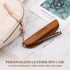 Puede incluir: Un estuche de cuero color canela con el nombre JASPER grabado en el frente, que contiene un bolígrafo de madera oscura con detalles metálicos. El estuche está unido a un bolso de lona crema mediante una cadena plateada y mide 19,8 cm L x 4 cm A.