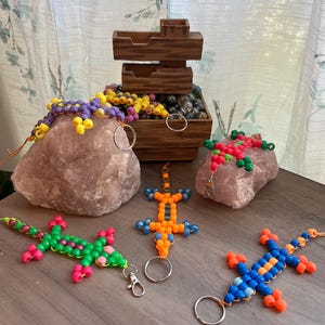 Puede incluir: Una colección de llaveros y pulseras de lagartos hechos con cuentas de colores. Los lagartos están hechos con cuentas de varios colores, incluyendo verde, rosa, naranja y azul. También hay una caja de madera con cuentas decorativas y una roca rosa.