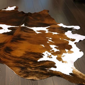 May include: A cowhide rug with a natural pattern of brown, white, and dark brown markings. The rug is irregularly shaped and lies on a dark wood floor, showcasing its unique texture and color variations. This decorative rug adds a rustic touch to any room.