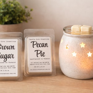 Op de afbeelding: Een brandende kaarsenwarmer met steruitsparingen naast twee wassmeltverpakkingen. De verpakkingen zijn gelabeld met "Brown Sugar" en "Pecan Pie". De warmer heeft een glazen bovenkant met twee wasblokjes.