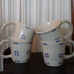 May include: A set of three ceramic mugs with a speckled, matte finish. Each mug features a different color: light blue, cream, and gray. The mugs have a simple, modern design and a comfortable handle.