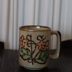 May include: A beige ceramic mug with a floral design. The mug features orange flowers, green leaves, and brown outlines. The handle is brown. The mug is on a dark wooden surface.