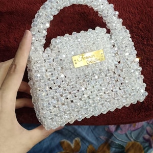 May include: A small handbag constructed entirely from clear, faceted, translucent beads. The bag features a rounded top handle and a rectangular gold-toned metal plaque on the front with the engraved text Farina's Collection in black script.