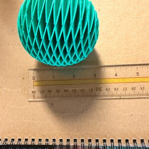 May include: A teal, 3D-printed sphere with a lattice design, approximately 3 inches in diameter, rests on a notebook. The sphere's intricate pattern creates a visually interesting texture. A ruler is placed next to the sphere for scale.