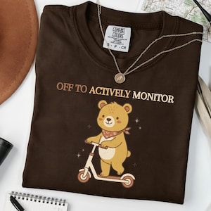 Può includere: Una maglietta marrone con la scritta "OFF TO ACTIVELY MONITOR" e un orso dei cartoni animati su uno scooter. L'orso indossa una bandana rossa. La maglietta ha un'etichetta Comfort Colors.