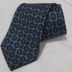 Puede incluir: Una corbata turquesa con un patrón repetitivo de medallones florales morados y azul marino. La corbata está doblada sobre sí misma, mostrando el intrincado diseño. La tela tiene una sutil textura acanalada, agregando profundidad.