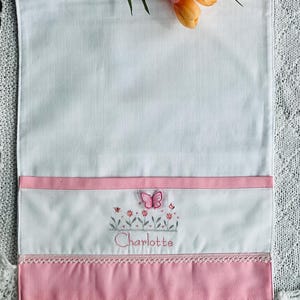 May include: A white rectangular fabric piece featuring a pink horizontal band at the bottom with a decorative lace trim. The center displays an embroidered design of small flowers, a pink butterfly, and the name Charlotte in pink cursive script.