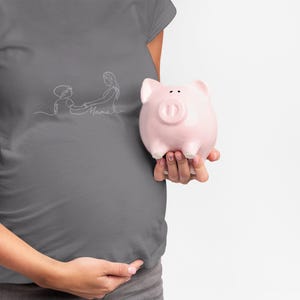 Op de afbeelding: Een grijs T-shirt met een witte lijntekening van een moeder en kind, met het woord "Mama" eronder. Een roze spaarvarken wordt in de hand gehouden. De afbeelding staat tegen een witte achtergrond.