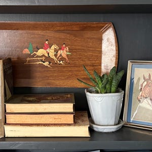 May include: A wooden serving tray with a hunting scene, books, a framed horse portrait, and a potted plant are displayed on a black shelf. The tray features riders on horseback and a dog.