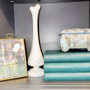 May include: A still life arrangement featuring a framed painting, a tall white vase, a small blue and brown decorative box, and three teal-colored books. The vase has a textured base and a flared top. The books are stacked.