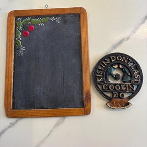 May include: A wooden-framed chalkboard with painted green leaves and red cherries in the corner, next to a black cast iron sign that reads "Kissin' Don't Last Cookin' Do."
