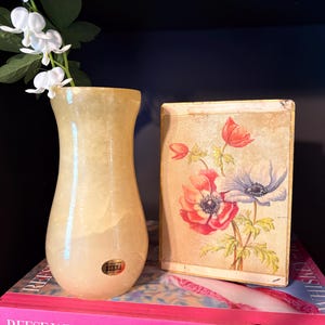 May include: A beige-colored vase with a flared top and a small label. The vase holds white flowers with green leaves. Beside the vase is a rectangular box with a floral design in red, pink, and blue.