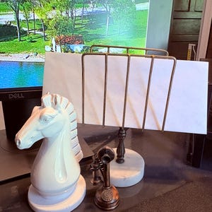 May include: A white ceramic chess knight figurine, a bronze-colored miniature telephone, and a brass and marble letter holder are displayed on a desk. The letter holder has a white paper inside.