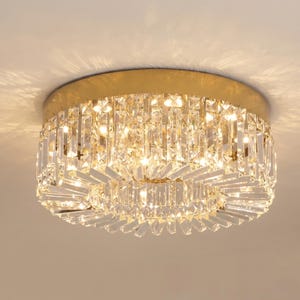 Peut inclure: Un plafonnier rond avec une base dorée et un abat-jour en cristal à double couche. Les cristaux sont rectangulaires et disposés en cercles concentriques, créant un effet scintillant. La lumière est allumée, illuminant les cristaux. Diamètre d'environ 30 cm.