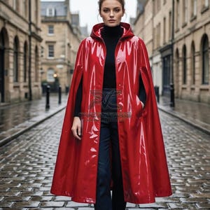 May include: A glossy red vinyl hooded cape featuring a front button closure and side arm slits. The garment is worn over a black turtleneck sweater and dark trousers. A logo with the text PVC DREAMS is visible on the front of the red cape.