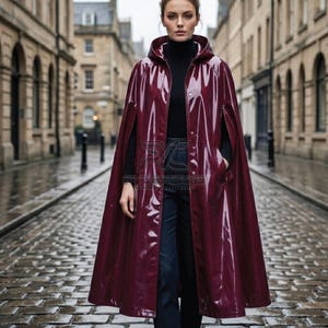 Könnte beinhalten: Burgunderfarbener Kapuzenmantel mit glänzendem Finish. Der Mantel hat eine Knopfleiste, Taschen und ein modernes Design. Das Model trägt einen schwarzen Rollkragenpullover und dunkle Hosen. Im Hintergrund eine Kopfsteinpflasterstraße.