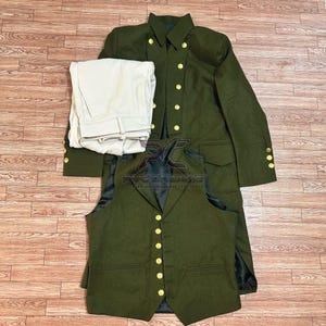May include: An olive green military-style uniform set, including a long coat with gold buttons, a vest, and cream-colored pants. The coat and vest have multiple gold buttons. The uniform is neatly arranged on a wooden surface.