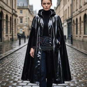May include: A glossy black vinyl hooded cape featuring a front button closure and arm slits. The garment has a high-shine finish and a structured hood. A logo with the text PVC DREAMS is visible on the chest area of the black vinyl material.