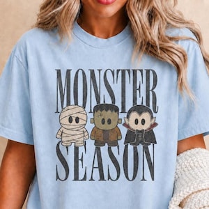 May include: A light blue short-sleeve t-shirt featuring the text MONSTER SEASON in a distressed black font. Between the words are three cartoon characters: a white mummy, a green Frankenstein-style monster, and a vampire in a black cape.