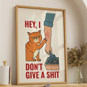 May include: A framed print featuring an orange tabby cat with an angry expression, clawing at an arm with scratches. The text reads "HEY, I DON'T GIVE A SHIT" in red. The cat is next to a litter box.