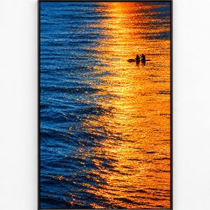 Può includere: Una stampa artistica da parete verticale incorniciata che raffigura uno specchio d'acqua calmo al tramonto. La superficie dell'acqua è divisa in ombre blu profondo a sinistra e vivaci riflessi di luce solare arancione a destra, con due sagome.