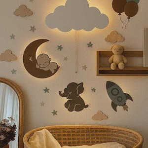 May include: A nursery wall decor scene featuring a cloud-shaped light fixture, a crescent moon with a sleeping baby, an elephant, a rocket, and star accents. A wicker bassinet and a teddy bear on a shelf complete the scene.