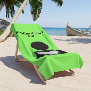 Może przedstawiać: Jasnozielony ręcznik plażowy w kolorze limonki z czarnym napisem Reading doesn't kill oraz stylizowaną czarno-białą grafiką przedstawiającą osobę czytającą książkę. Ręcznik jest rozłożony na drewnianym leżaku na piaszczystej plaży.