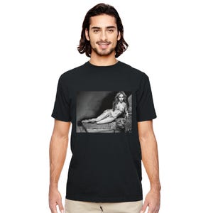 Könnte beinhalten: Schwarzes T-Shirt mit einem Schwarz-Weiß-Bild einer Person, die auf einer Steinoberfläche liegt. Das Bild ist eine detaillierte Zeichnung oder ein Foto, wobei der Fokus auf der Pose der Person und den Texturen von Stein und Kleidung liegt.