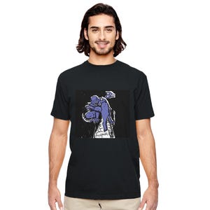Può includere: T-shirt nero con una grafica di una figura blu in stile usurato. La figura è accovacciata su una struttura di mattoni bianchi. La grafica è di forma rettangolare.